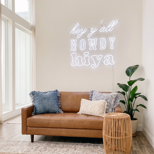 Neon sign on a wall above a brown leather sofa with decorative pillows and a plant in a room with large windows.