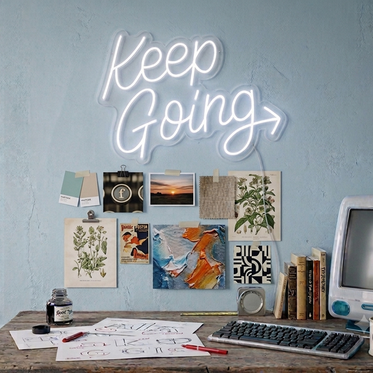 Neon sign reading 'Keep Going' above a desk with various items and wall art.