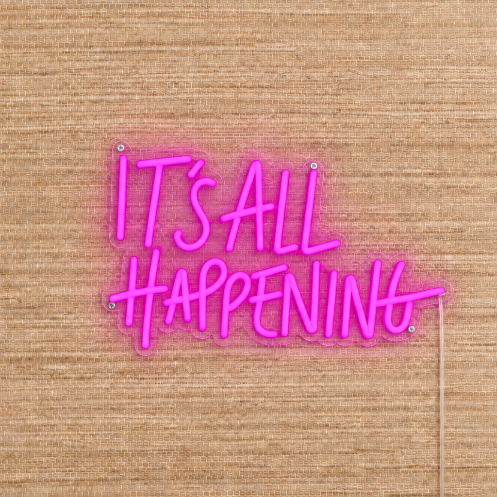 Flamingo Pink It’s All Happening LED neon sign on beige woven wallpaper with clear acrylic backing and silver mounting screws.