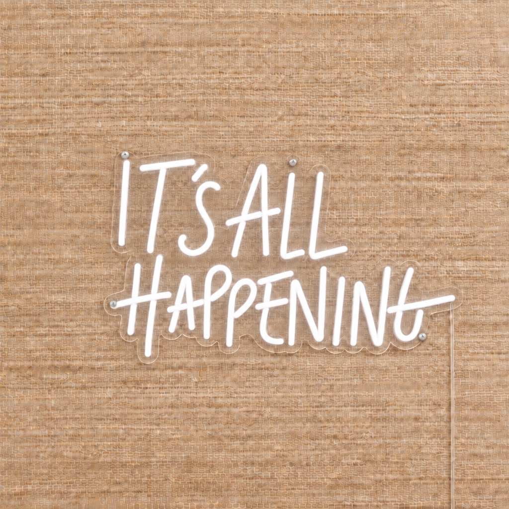 White It’s All Happening LED neon sign on beige woven wallpaper with clear acrylic backing and silver mounting screws.